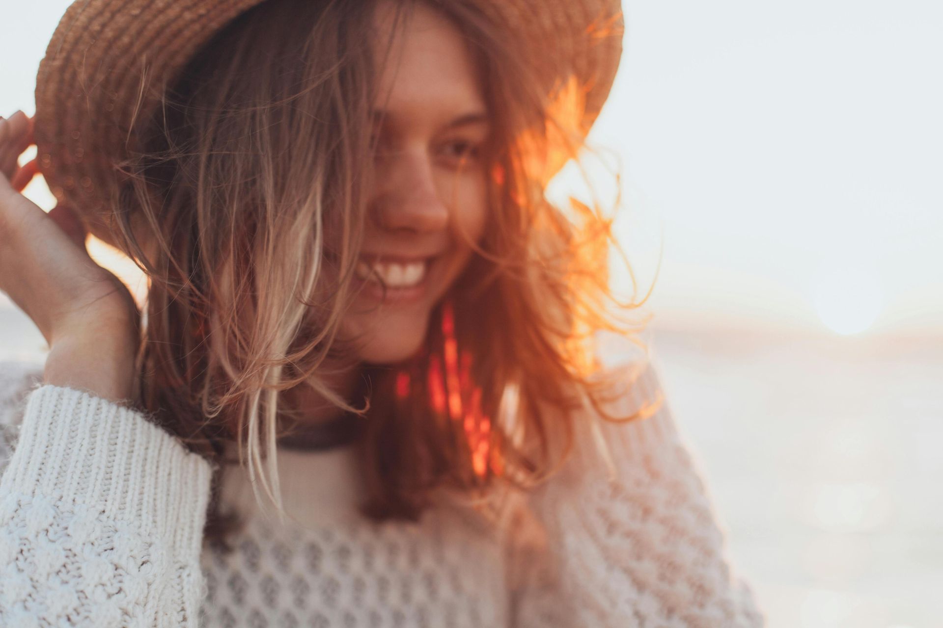 Joyful woman in a straw hat smiling by the seaside with sunset glow.